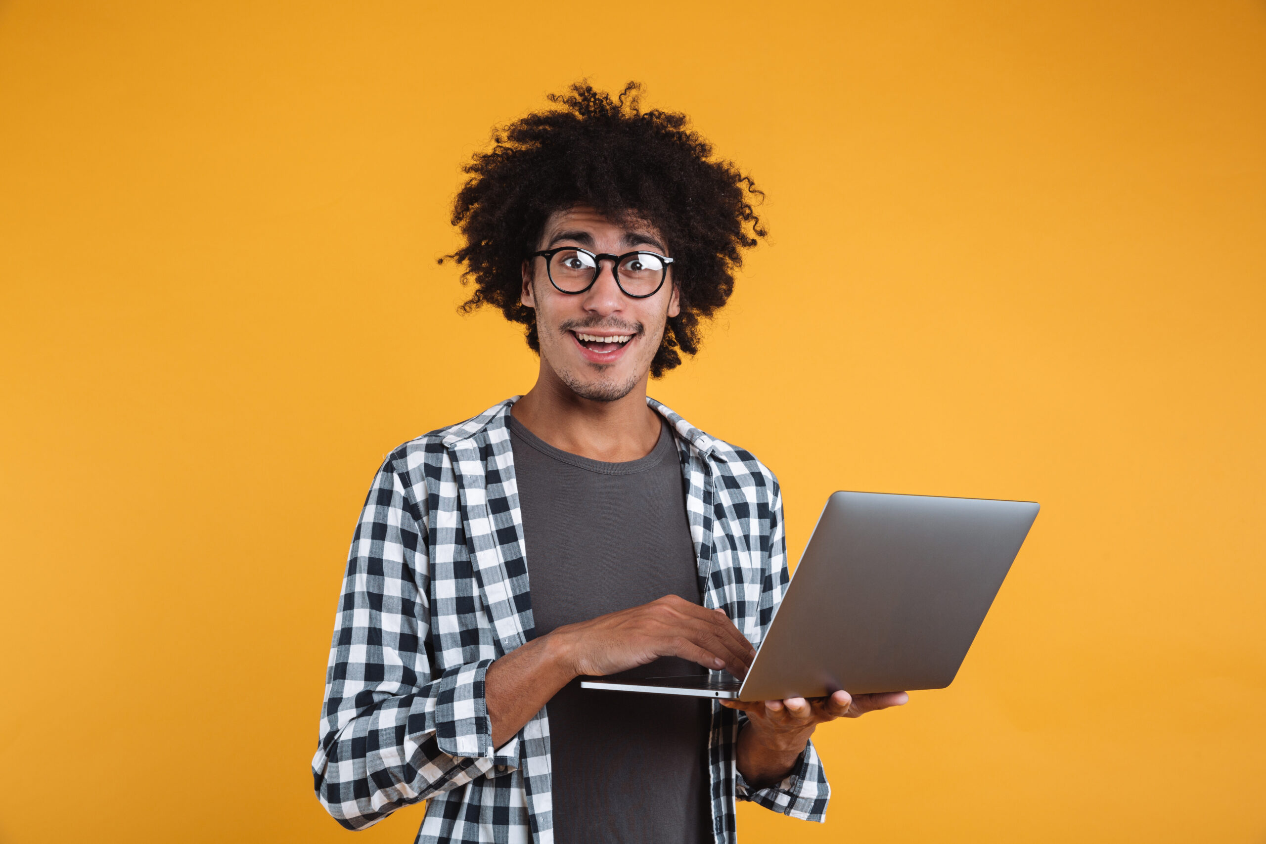 Portrait of a happy young african man in eyeglasses holding laptop computer and looking at camera isolated over orange background